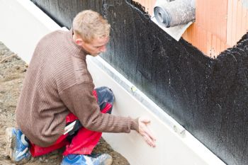 Worker applying waterproofing coating to an exterior foundation wall near a house base